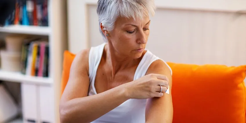 Woman with gray hair applying a hormone therapy patch to her upper arm at home